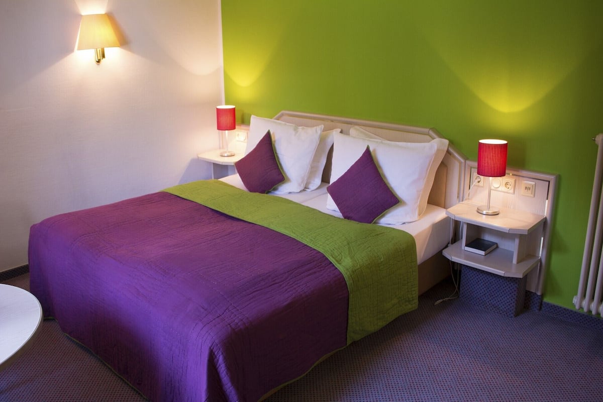Une chambre d’hôtel comprenant un lit, deux tables de chevet, une petite table, une chaise et un mur d’accent vert.