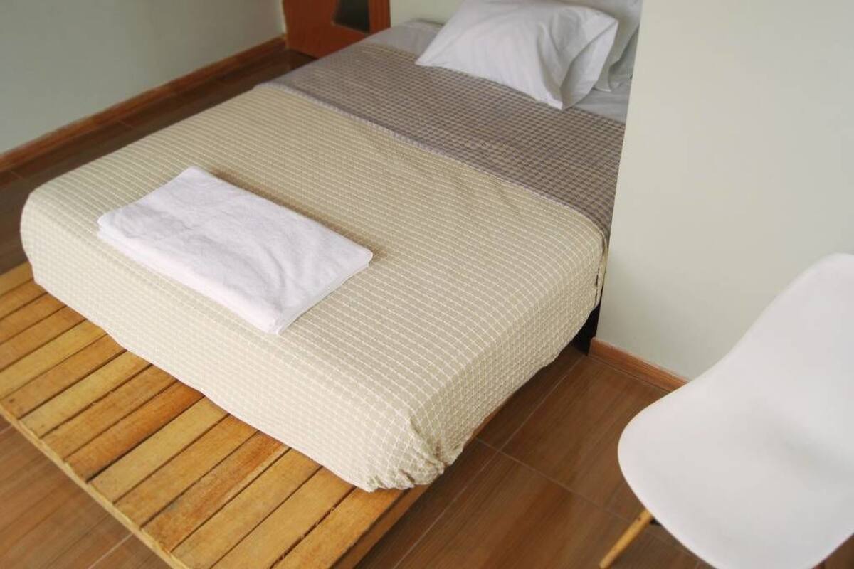 Ein ordentlich bezogenes Bett mit einem weißen Handtuch darauf, Holzboden und ein weißer Stuhl zur Rechten.