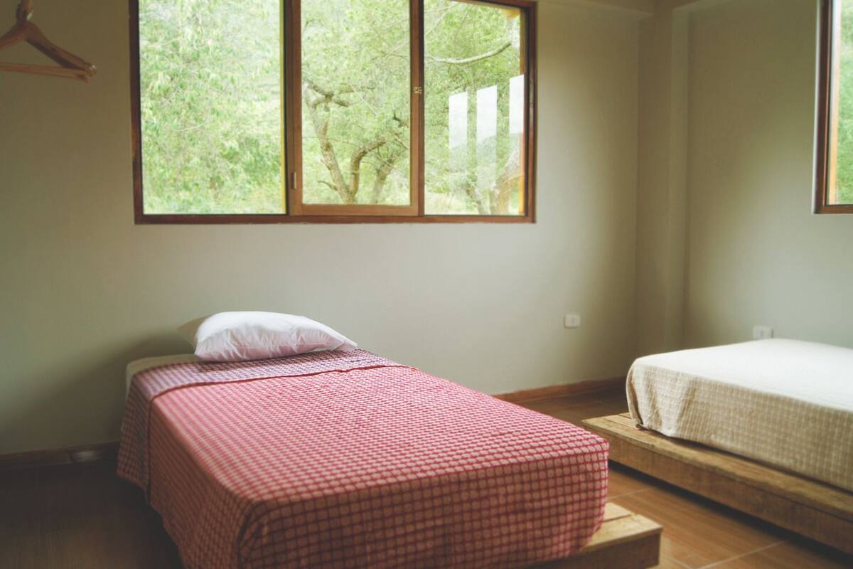 Ein Zimmer mit zwei Einzelbetten, eines mit einer rot karierten Bettdecke und das andere mit einer weißen. Ein Fenster bietet Blick auf Bäume.