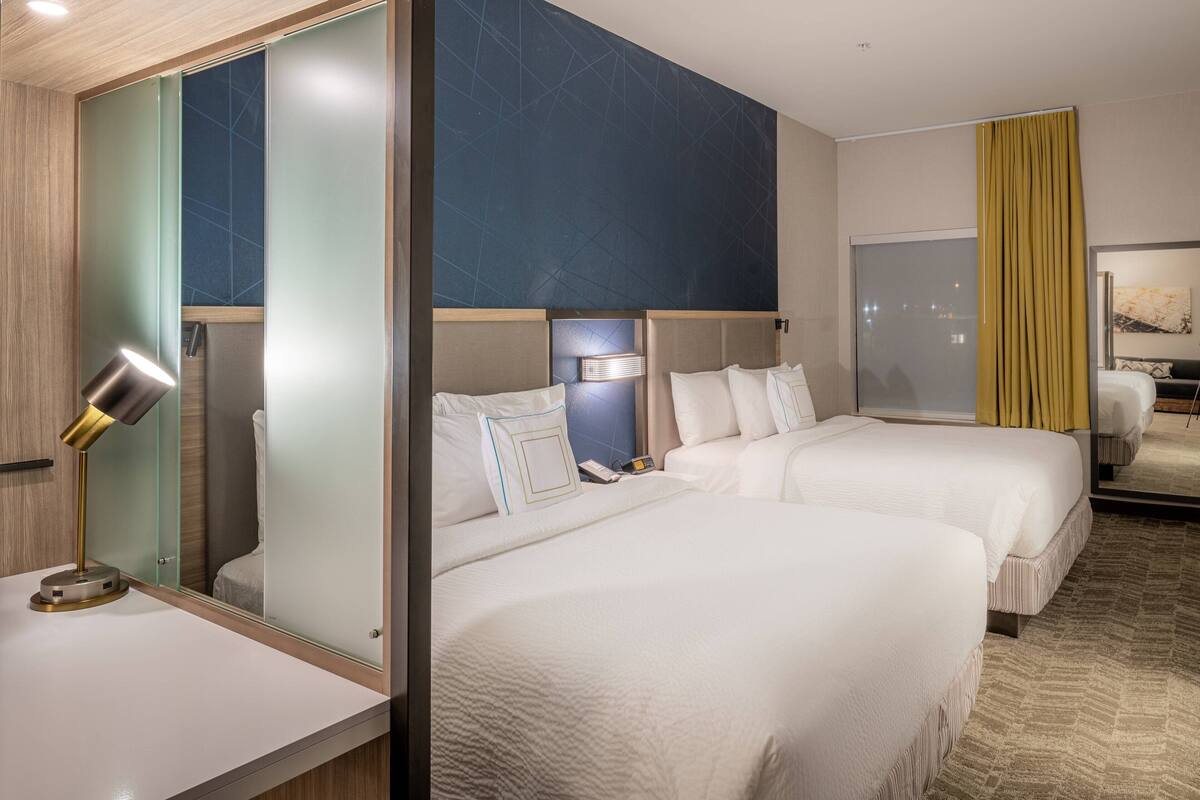A hotel room with two beds, a desk with a lamp, a mirror, and a large window with curtains.