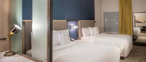 A hotel room with two beds, a desk with a lamp, a mirror, and a large window with curtains.