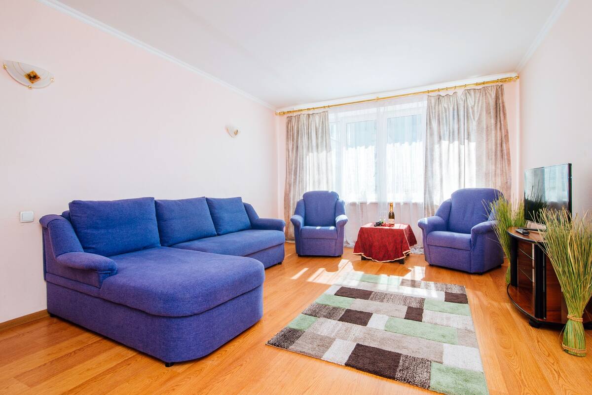 A living room with a blue sofa, two armchairs, a small table with a red tablecloth, and a TV stand with a vase of plants.
