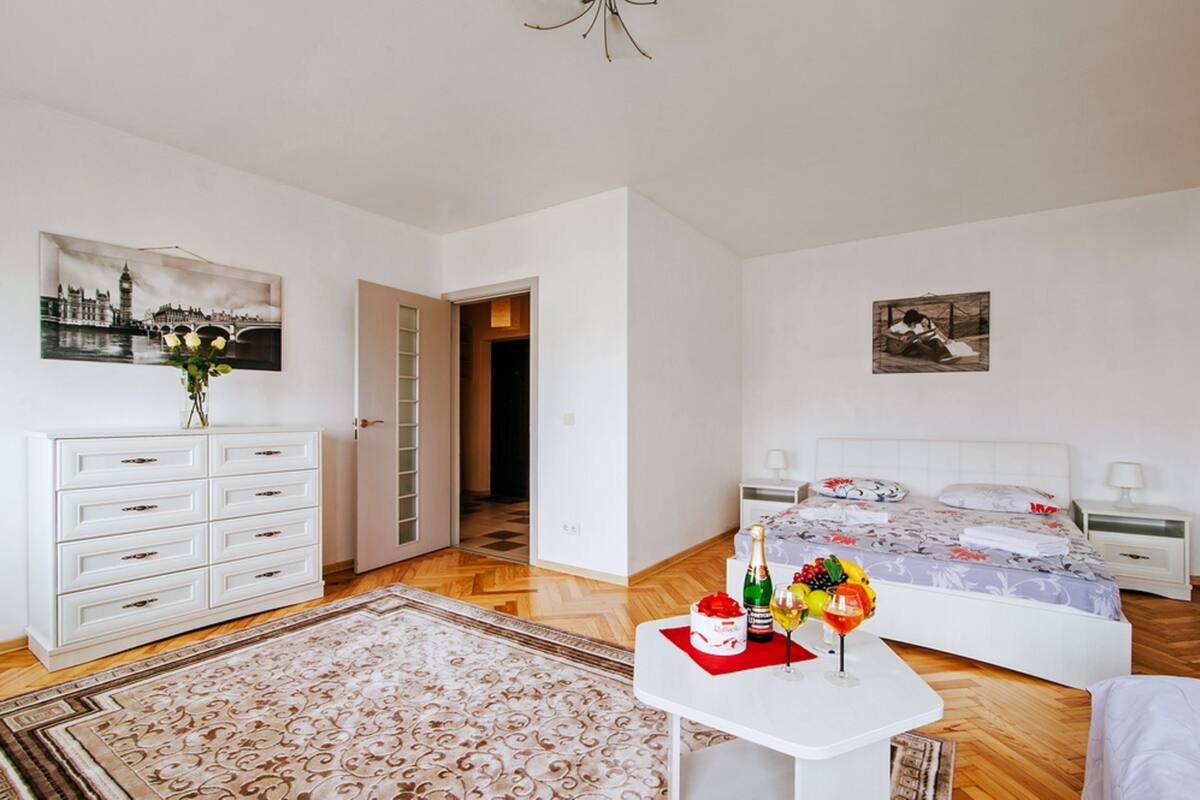 A hotel room with a bed, a nightstand, a bedside table with a bottle and flowers, a dresser, a chair, a door, and a hallway.