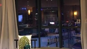 Une chambre d’hôtel avec une fenêtre incurvée, une vue sur la ville la nuit, un lit avec un oreiller vert et une petite table sur laquelle se trouve un sac vert.