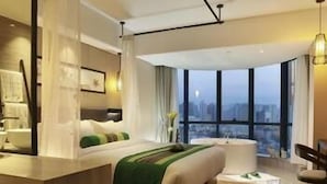 Une chambre d’hôtel moderne dotée d’un grand lit, d’un coin salon avec un fauteuil, d’un bureau et offrant une vue sur la ville grâce à de grandes fenêtres.