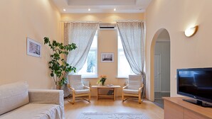 Un salon comprenant un canapé, deux fauteuils, une table basse ornée de fleurs, un téléviseur posé sur un meuble et une grande fenêtre avec des rideaux.