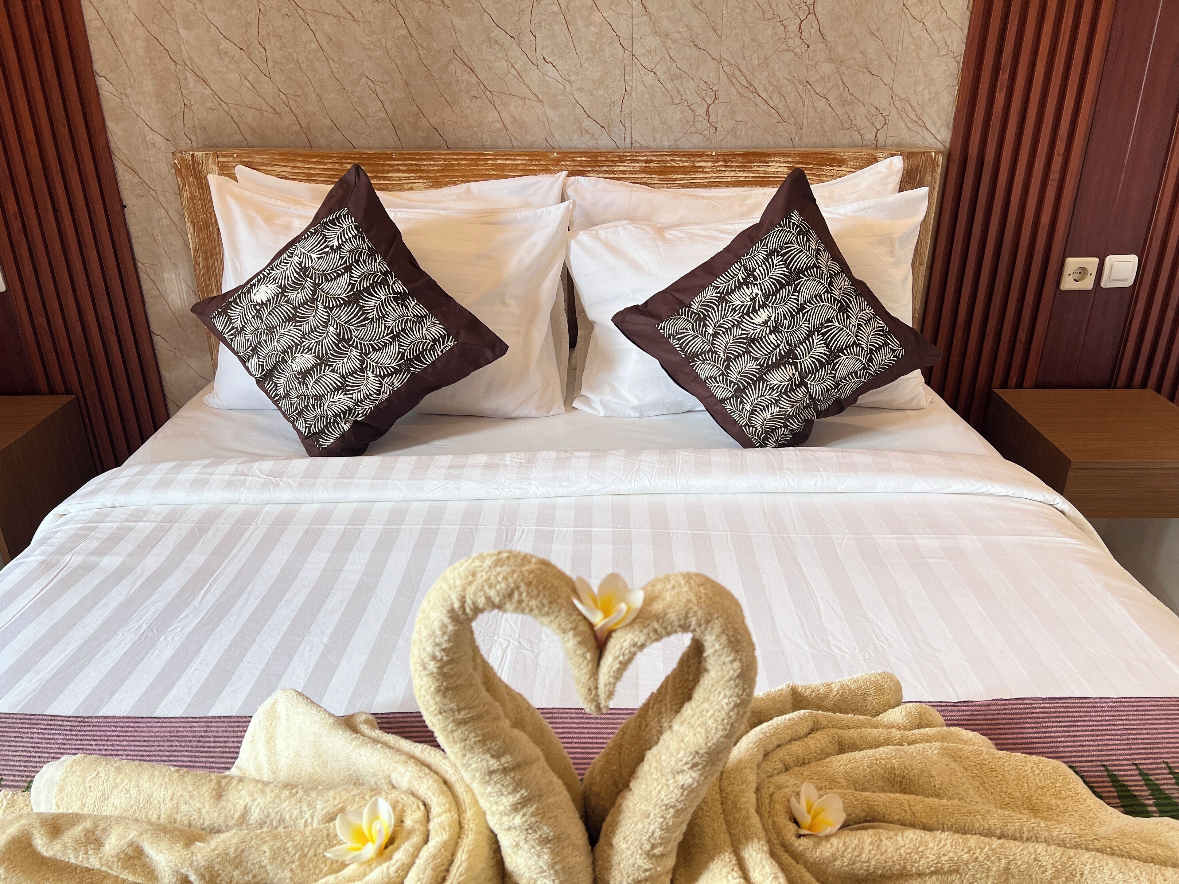 A neatly made bed with white linens, two decorative pillows with a leaf pattern, and two towels arranged in a heart shape with flowers.
