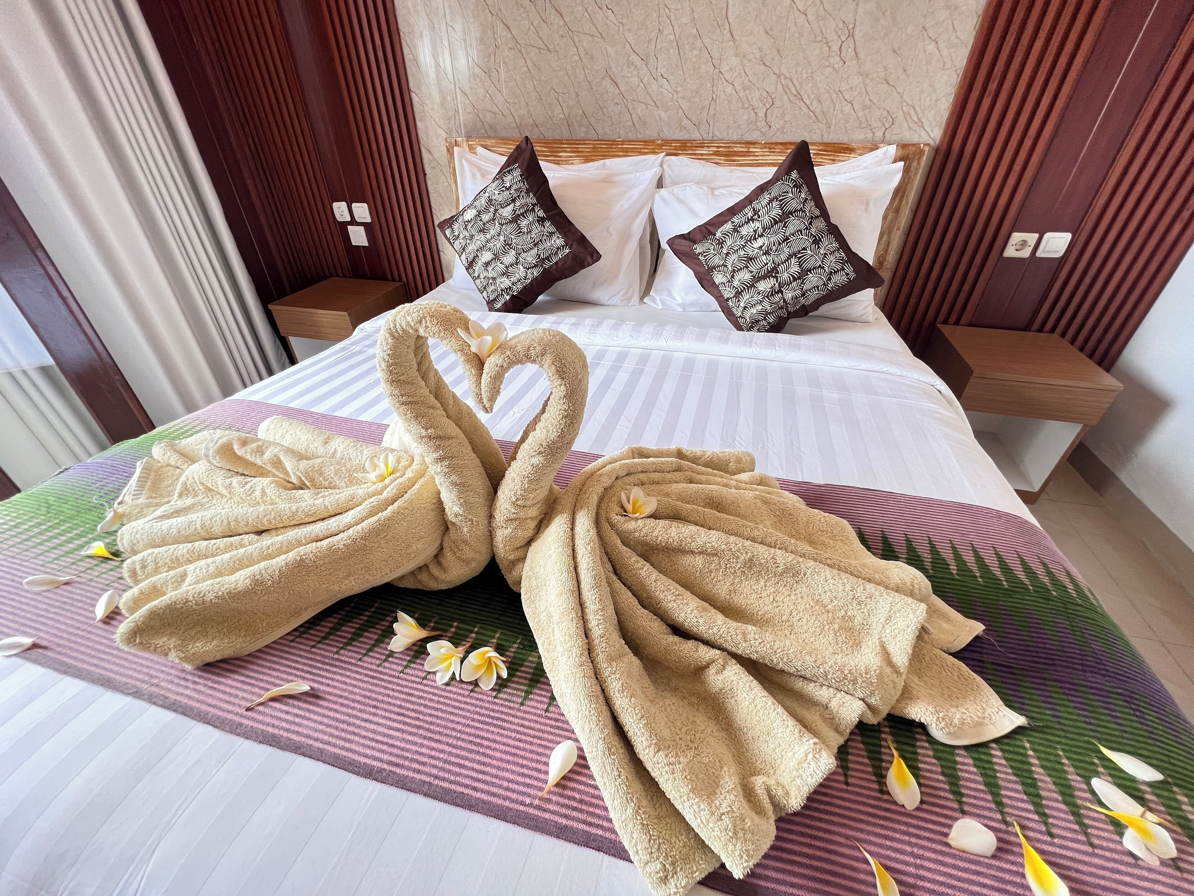A hotel room with a neatly made bed, two pillows, and a decorative towel arrangement on the bedspread.