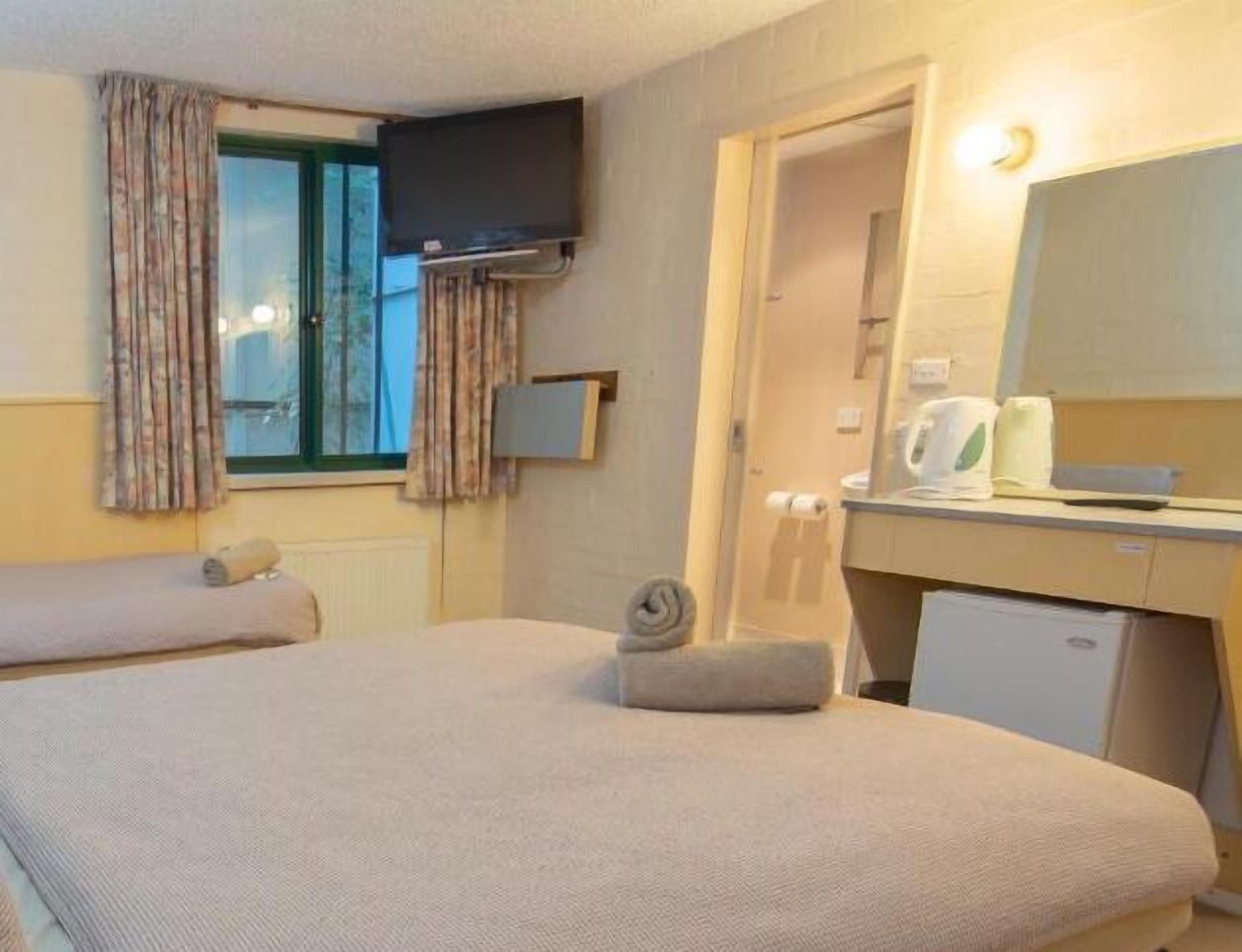 A hotel room with two beds, a TV mounted on the wall, a small desk with a mirror, and a window with curtains.