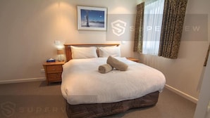 Quarto de hotel com cama grande, mesa de cabeceira, luminária, quadro na parede e janela com cortinas.