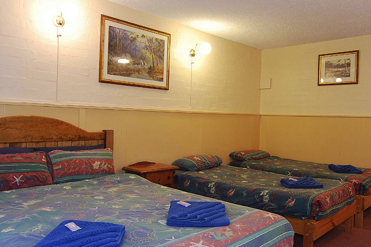 A room with three beds, each with a different patterned bedspread. There are two wall-mounted light fixtures and two framed pictures on the walls.