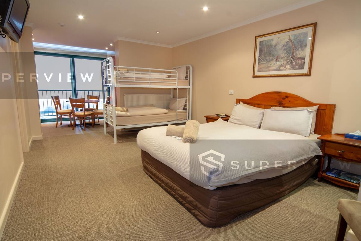A hotel room with a bunk bed, a single bed, a wooden headboard, a painting on the wall, a small table with a lamp, and a view of the outside through a window.