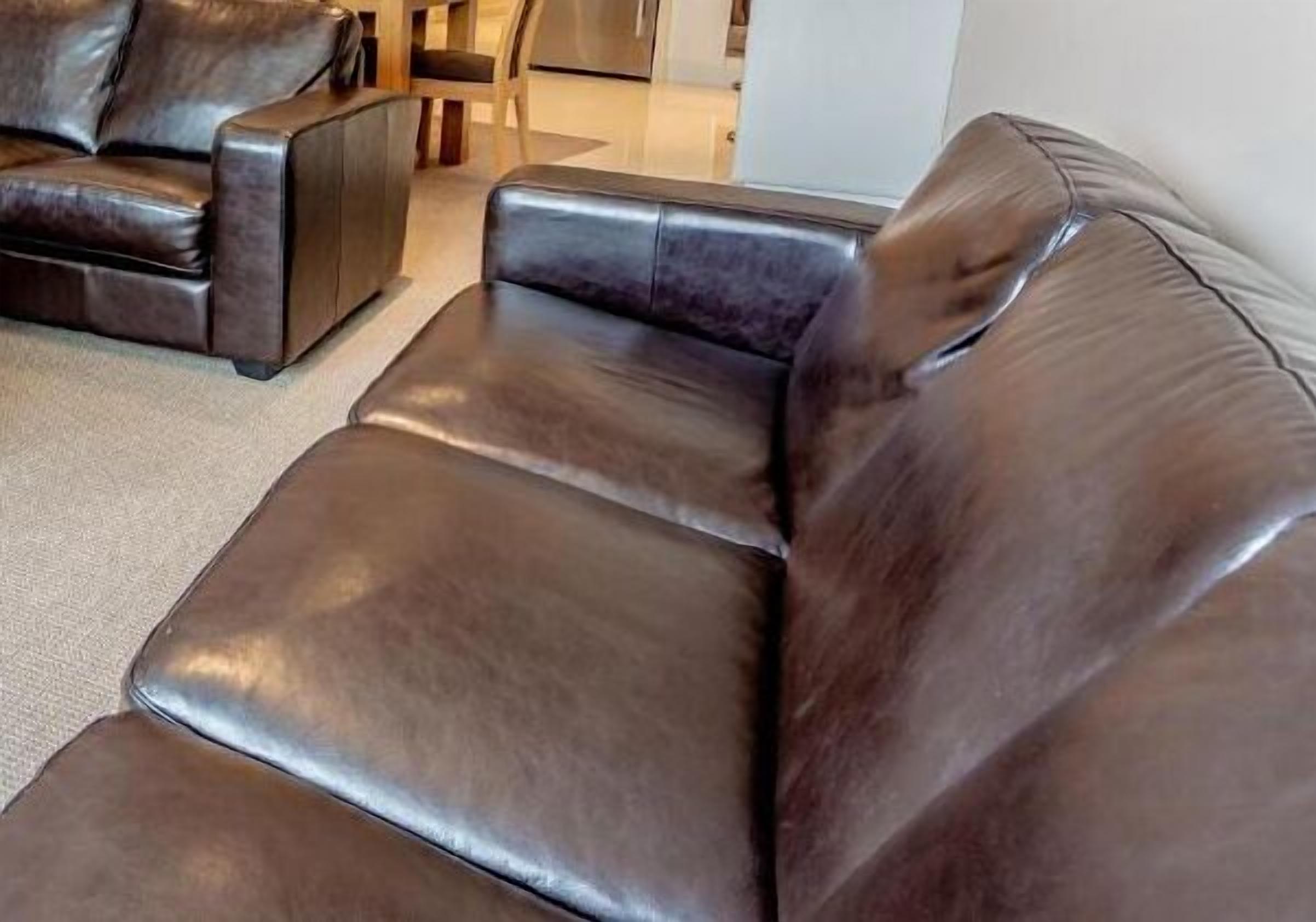 A dark brown leather sofa with a high backrest and armrests, positioned in a modern living room with a neutral color palette and minimalistic decor.