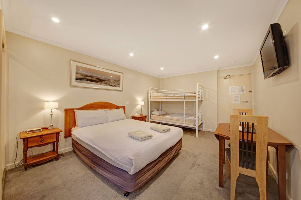 A bedroom with a single bed, a bunk bed, a desk, a chair, a TV, and a nightstand with a lamp.