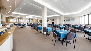 Uma sala de conferências ampla, com várias mesas e cadeiras, um balcão de recepção e vista para a cidade através de grandes janelas.