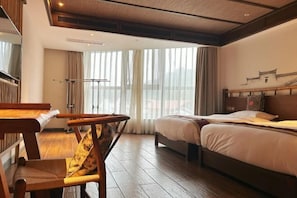 A hotel room with a wooden floor, a bed with white bedding, a wooden desk with a chair, and a large window with curtains.