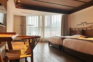 Quarto de hotel com piso de madeira, uma cama com roupa de cama branca, uma escrivaninha de madeira com uma cadeira e uma janela grande com cortinas.