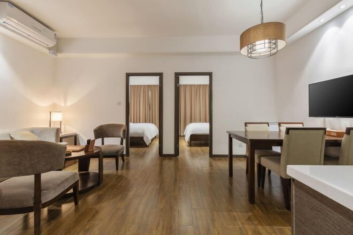A hotel room with a wooden floor, a sofa, two chairs, a dining table, a TV, and a large window with curtains.