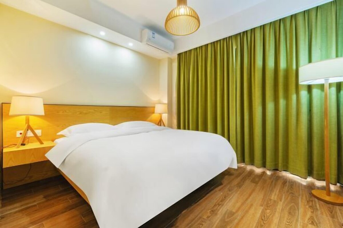 A hotel room with a bed, two bedside lamps, a wooden headboard, a green curtain, and a floor lamp.