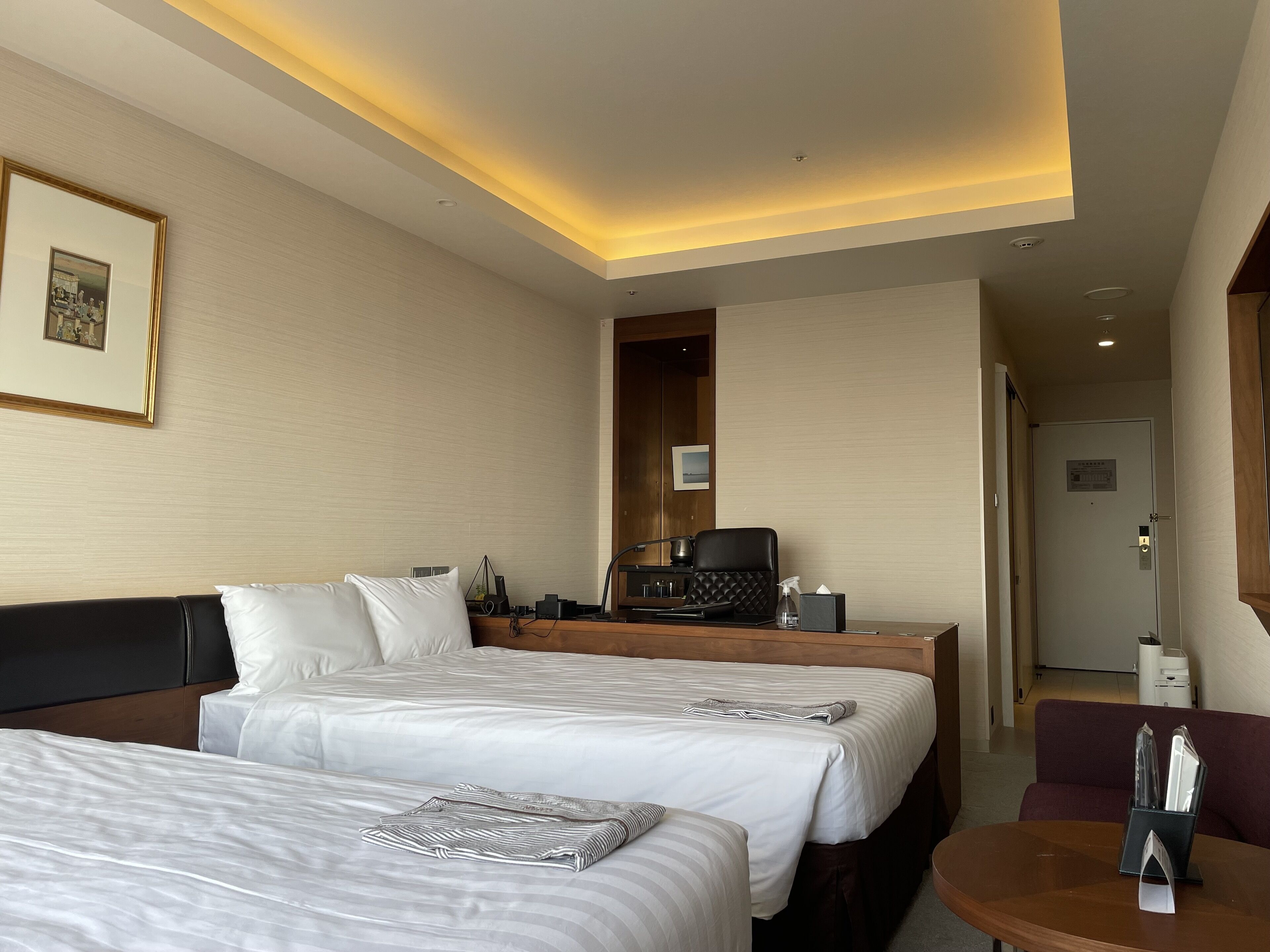 Habitación de hotel con dos camas, un escritorio con silla, una cafetera y una mesita con caja de pañuelos.