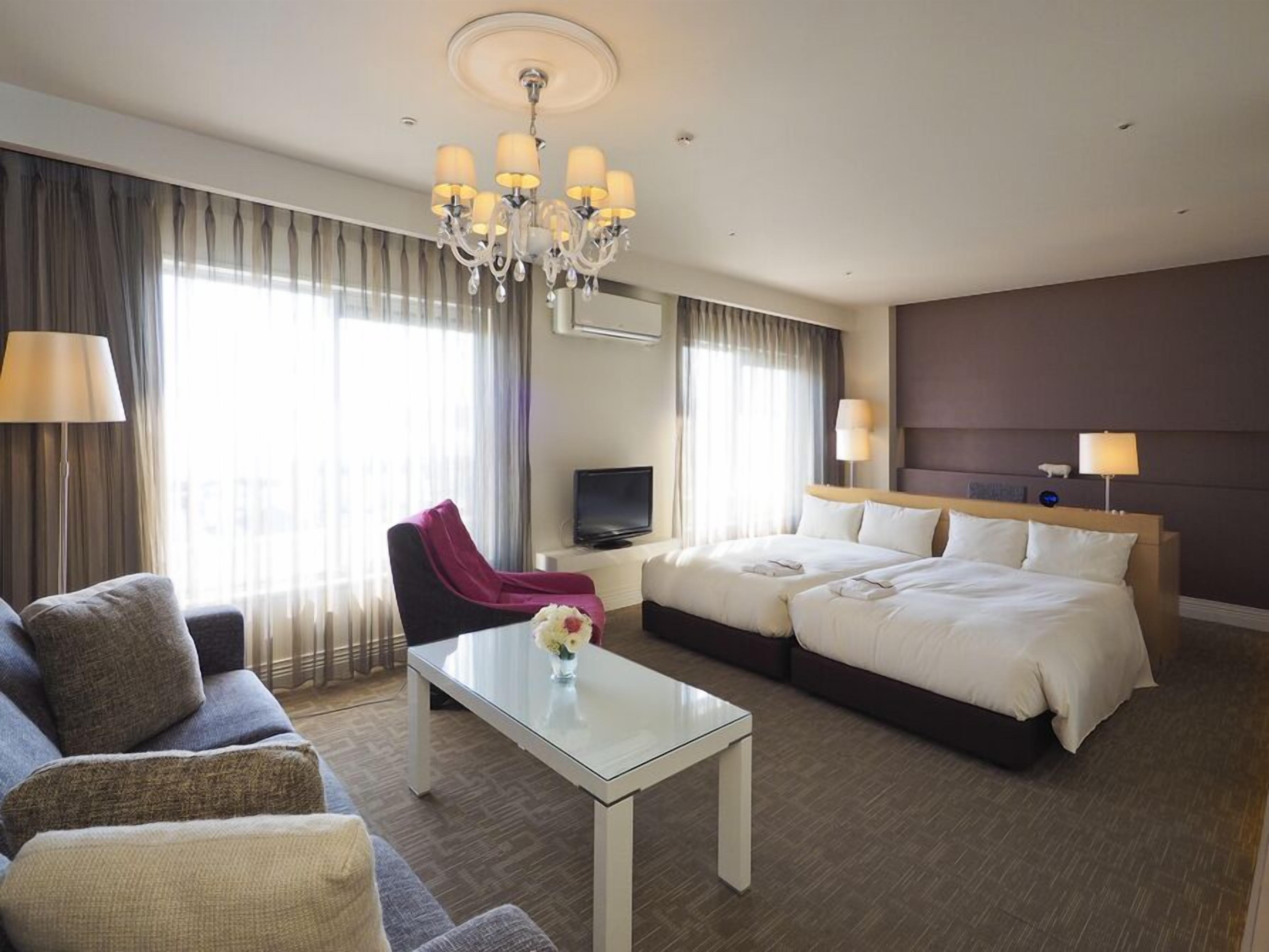 Une chambre d’hôtel comprenant un lit, deux tables de chevet, un canapé, une chaise, une télévision et un lustre.
