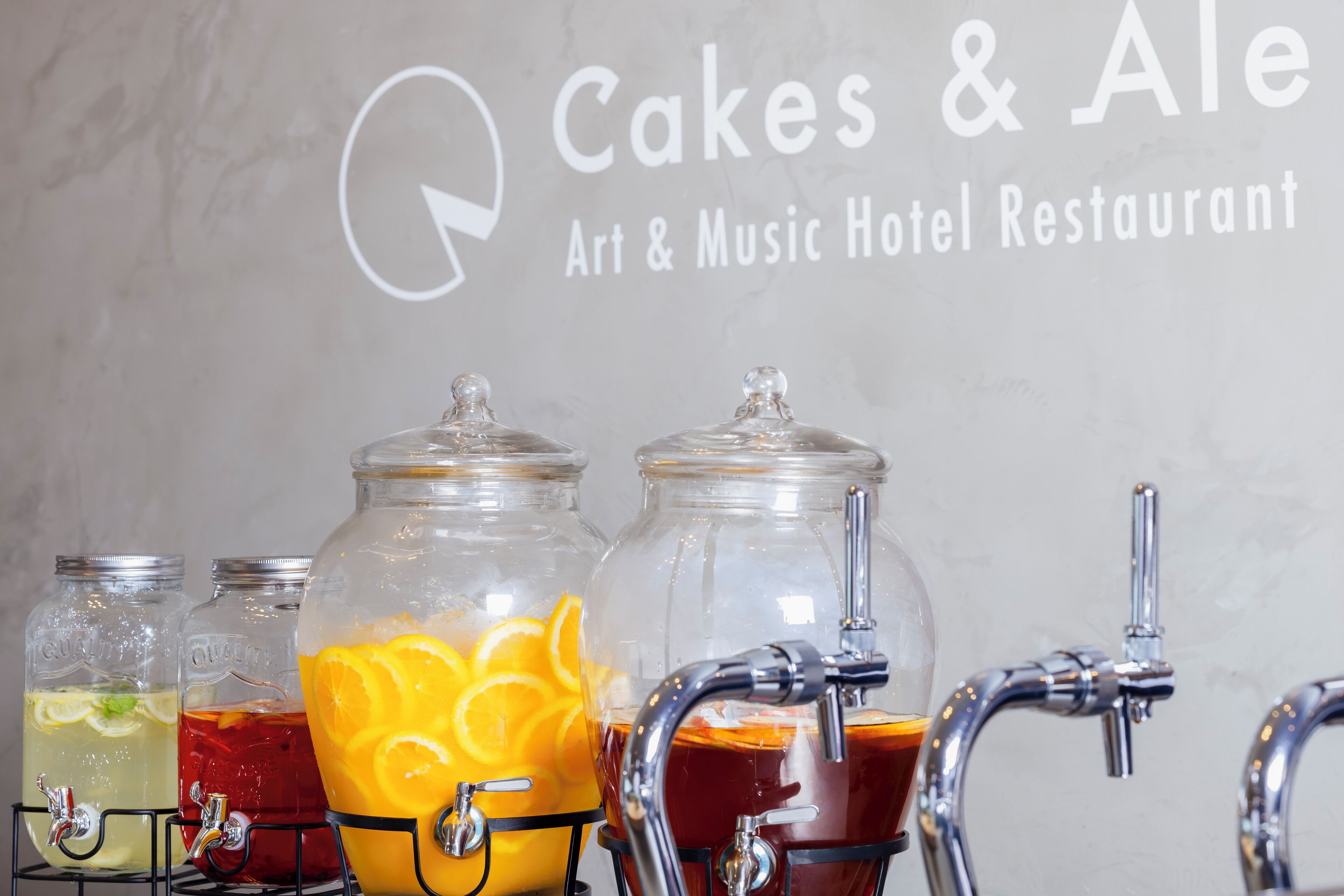 Una estación de bebidas con tres frascos de vidrio que contienen diferentes bebidas, un grifo y un letrero que dice 'Cakes & Ale Art & Music Hotel Restaurant'.