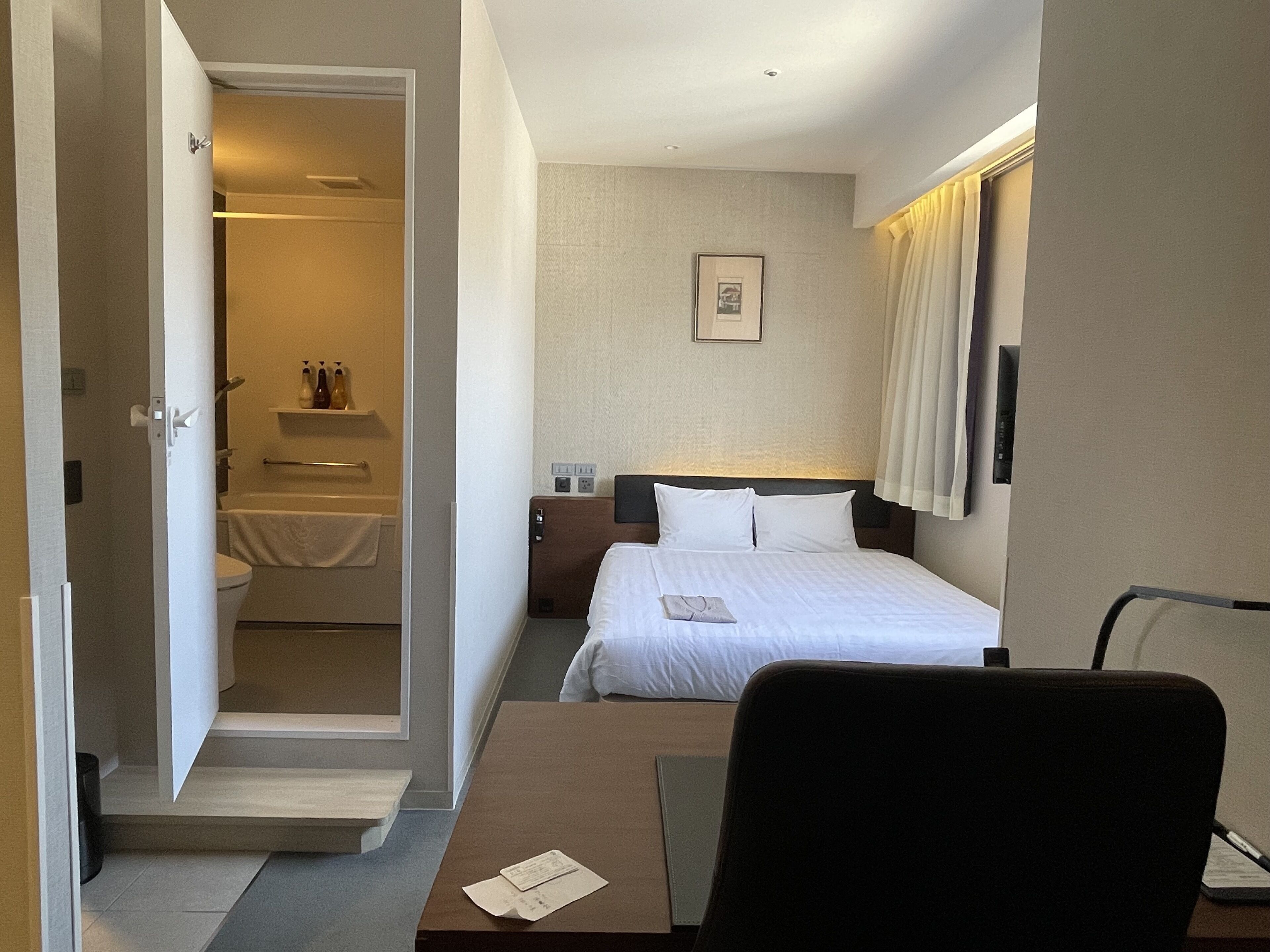 Ein Hotelzimmer mit Bett, Schreibtisch, Stuhl, Nachttisch, einem Bild an der Wand und einem durchgegangenen Tür erkennbaren Badezimmer.