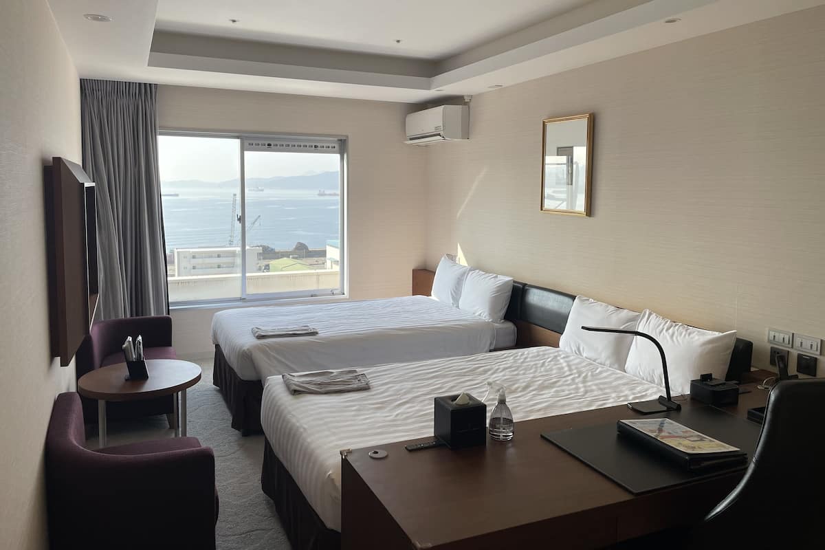 Een hotelkamer met twee bedden, een bureau, een stoel, een klein tafeltje en uitzicht op zee.