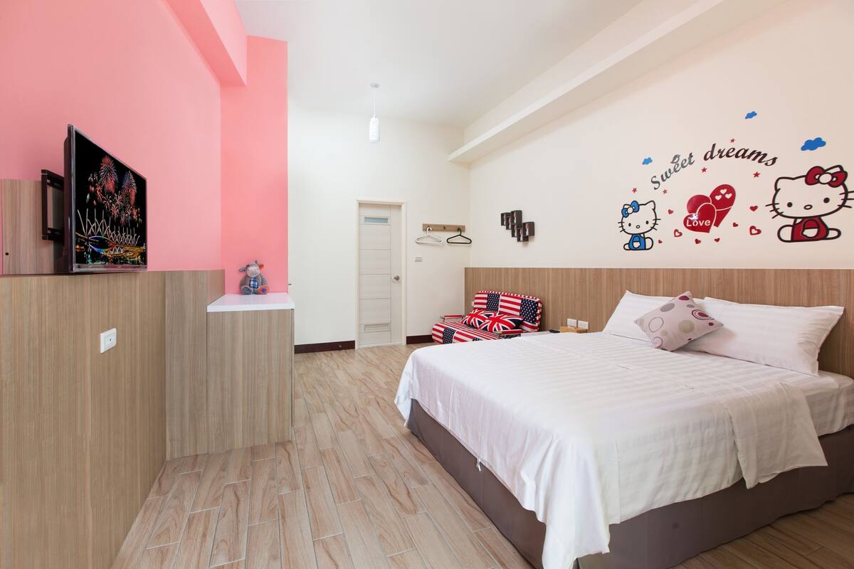 A bedroom with a bed, a TV on a stand, a door, and a wall with a Hello Kitty decoration.