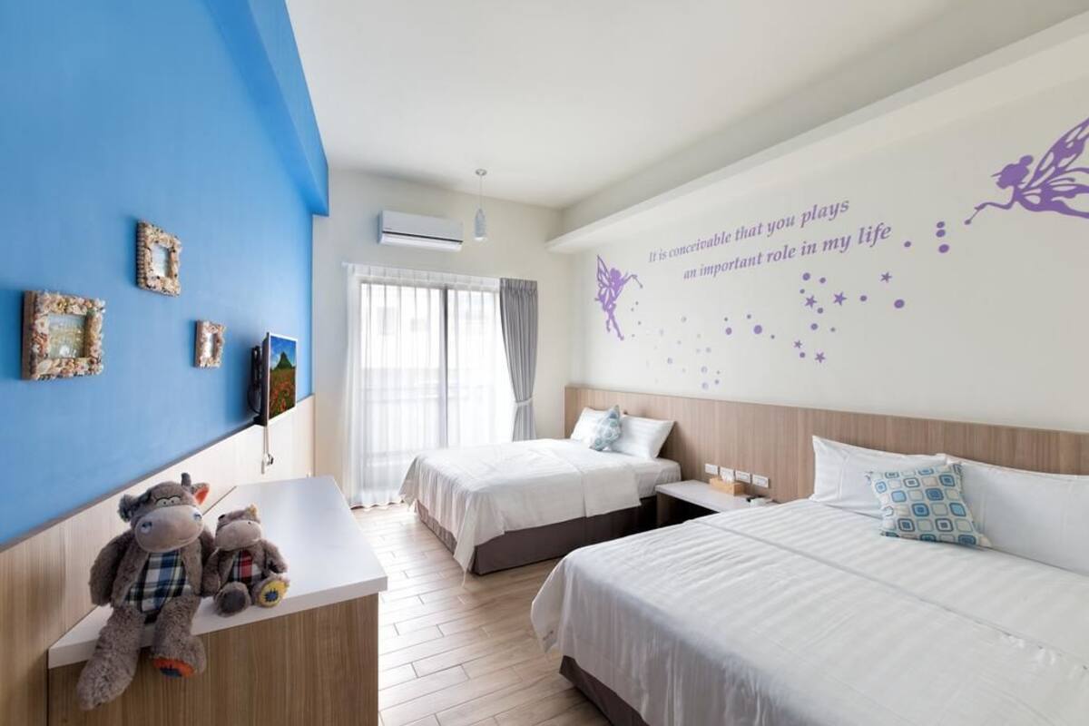 A hotel room with two beds, a desk with plush toys, a wall with a decorative quote, and a large window with curtains.