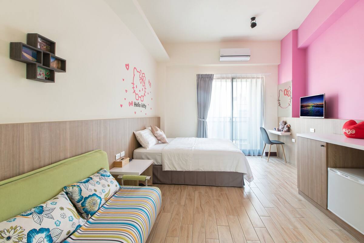 A modern hotel room with a bed, a desk, a small kitchenette, and a seating area with a sofa.