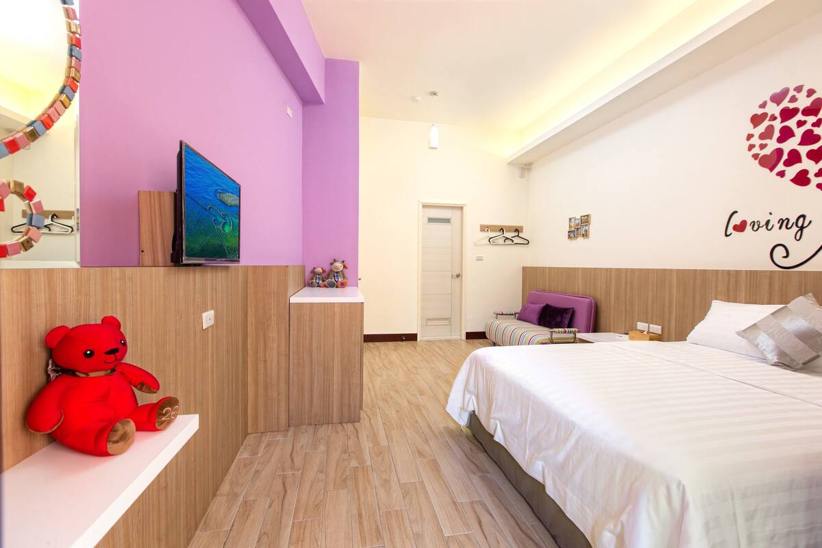 A bedroom with a bed, a TV on a shelf, a teddy bear, and a wall with a heart decoration.