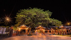 A traditional building with multiple rooms, a large tree in front, and a gated entrance.