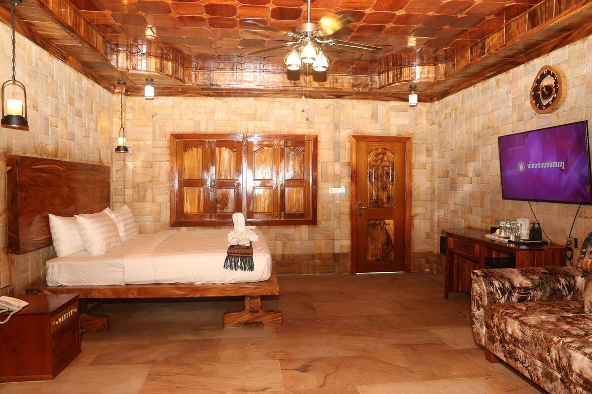 A room with a bed, wooden headboard, a TV, a wooden desk, and a stone wall.