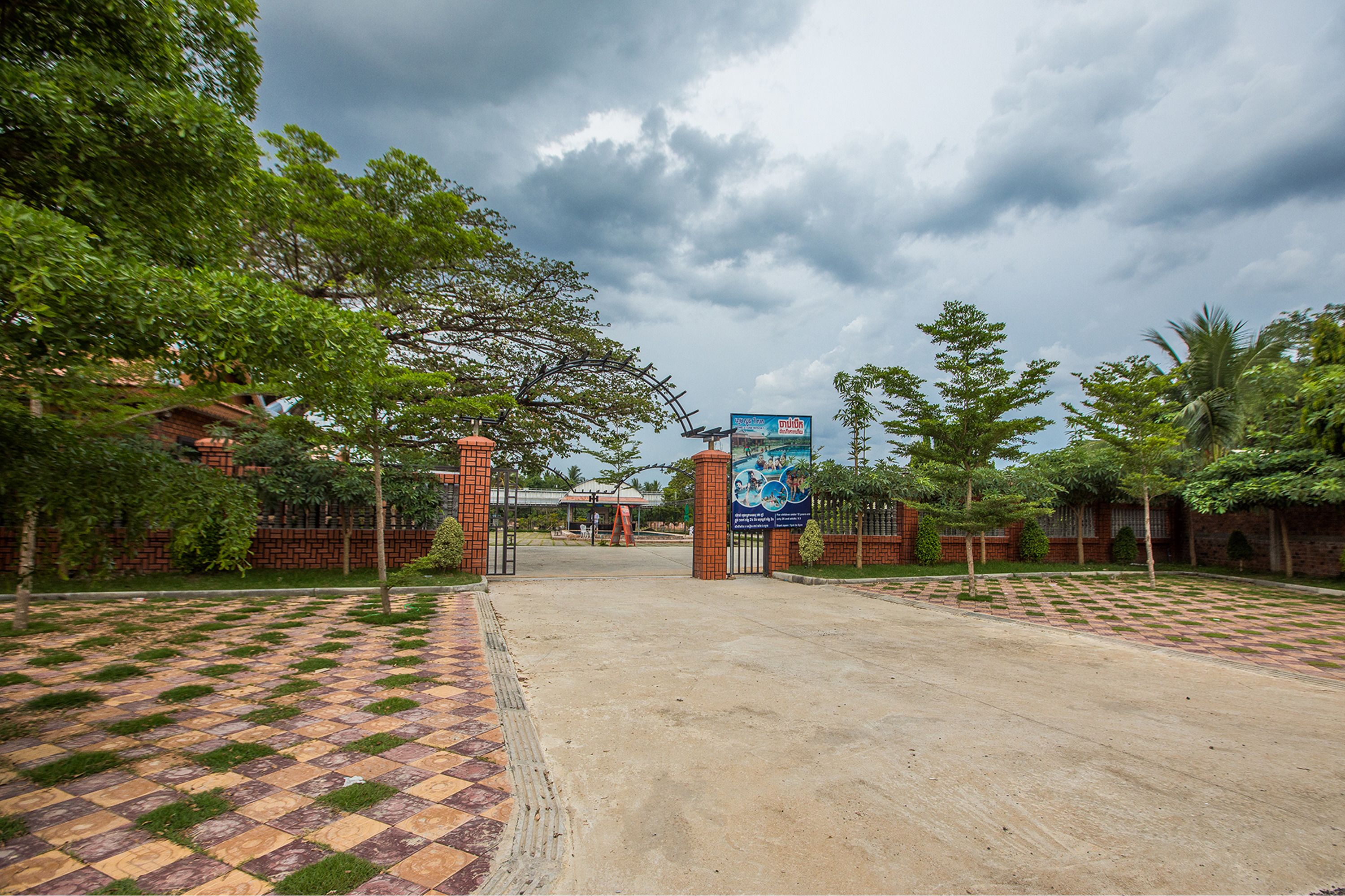 A gated entrance with a checkered pavement leading up to a building, surrounded by greenery and a cloudy sky.