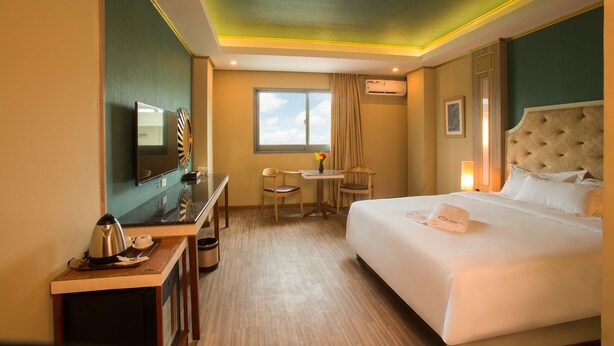 Een hotelkamer met een bed, een bureau met waterkoker, een stoel, een tafeltje met een vaas, en een raam met gordijnen.