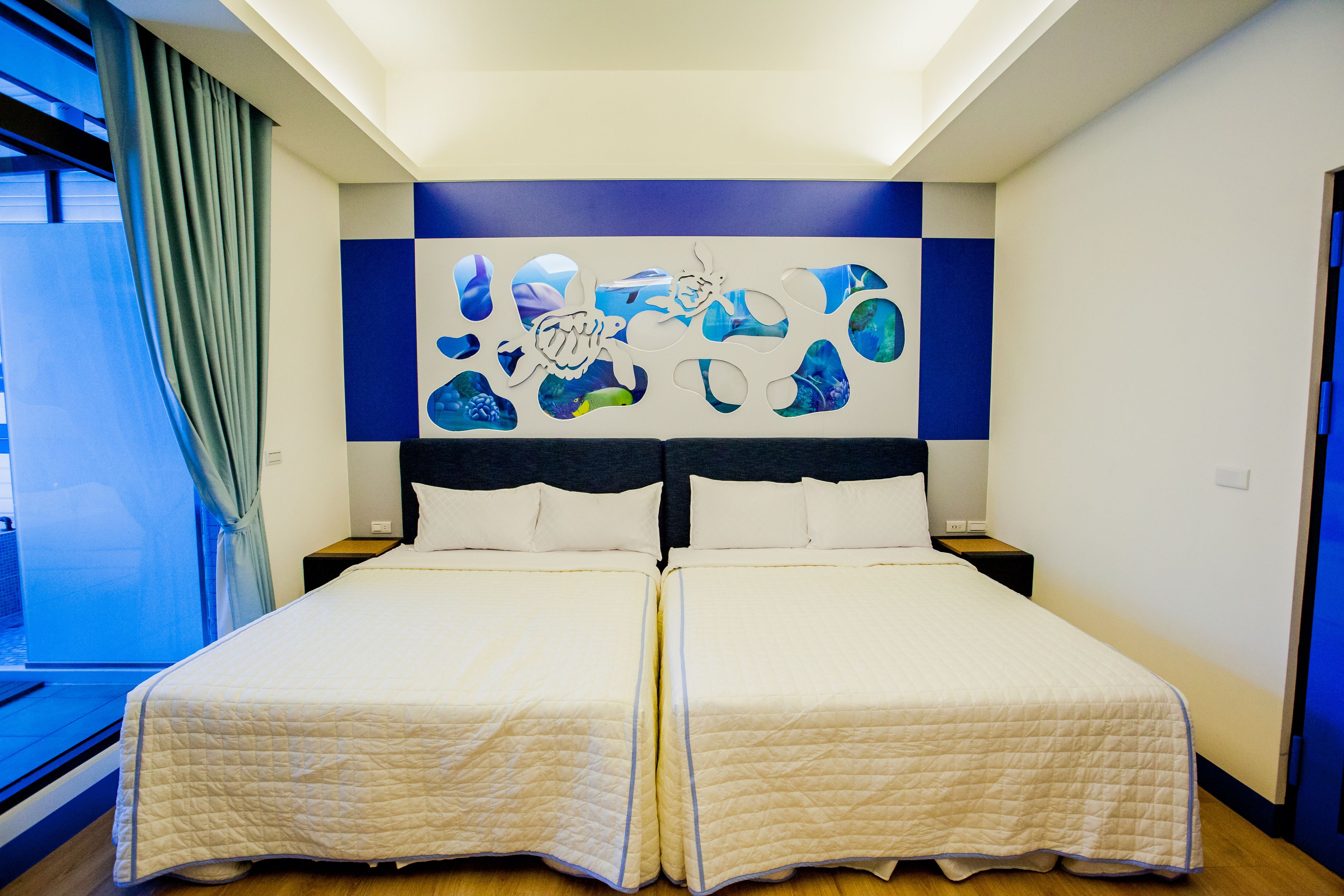A double bed with a quilted duvet, a headboard with a blue and white abstract design, and a window with a curtain.