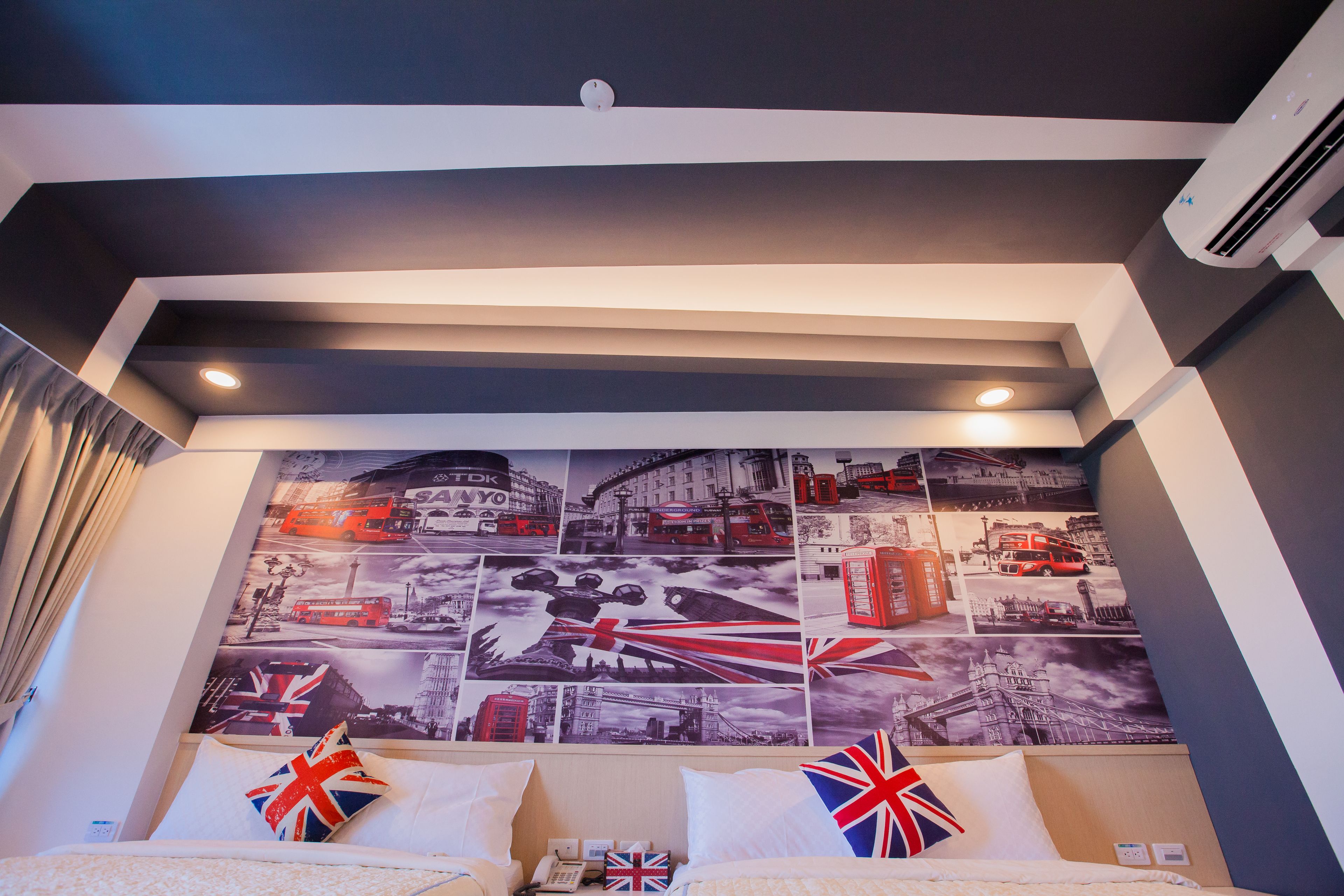A hotel room with a large wall mural of London landmarks, a bed with two pillows featuring the Union Jack, and an air conditioning unit mounted on the wall.