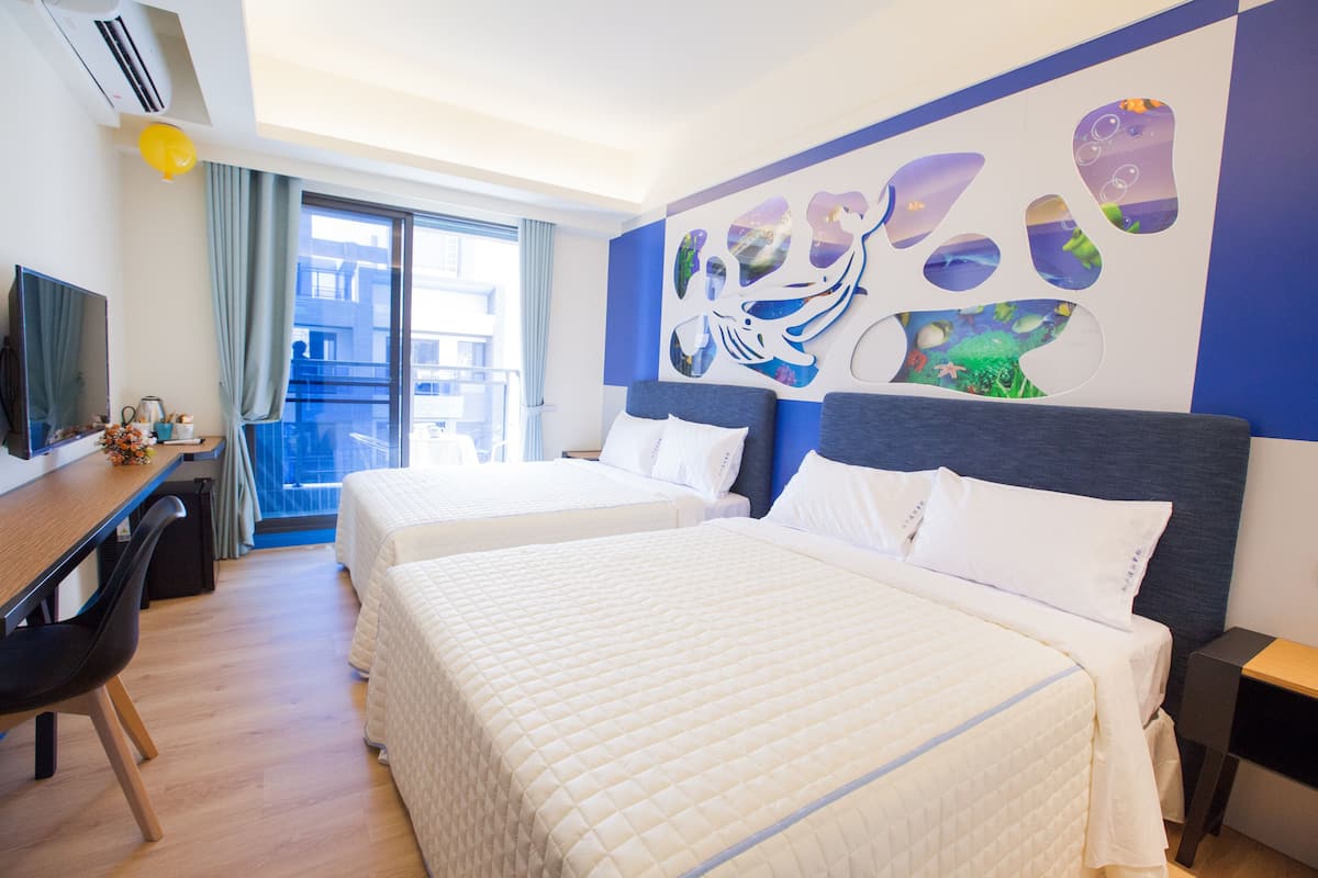 Habitación de hotel con dos camas, un escritorio, una silla y un ventanal grande.