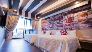 Habitación de hotel con dos camas, un ventanal grande con cortinas, una pared decorada con carteles de viajes y un televisor de pantalla plana montado en la pared.