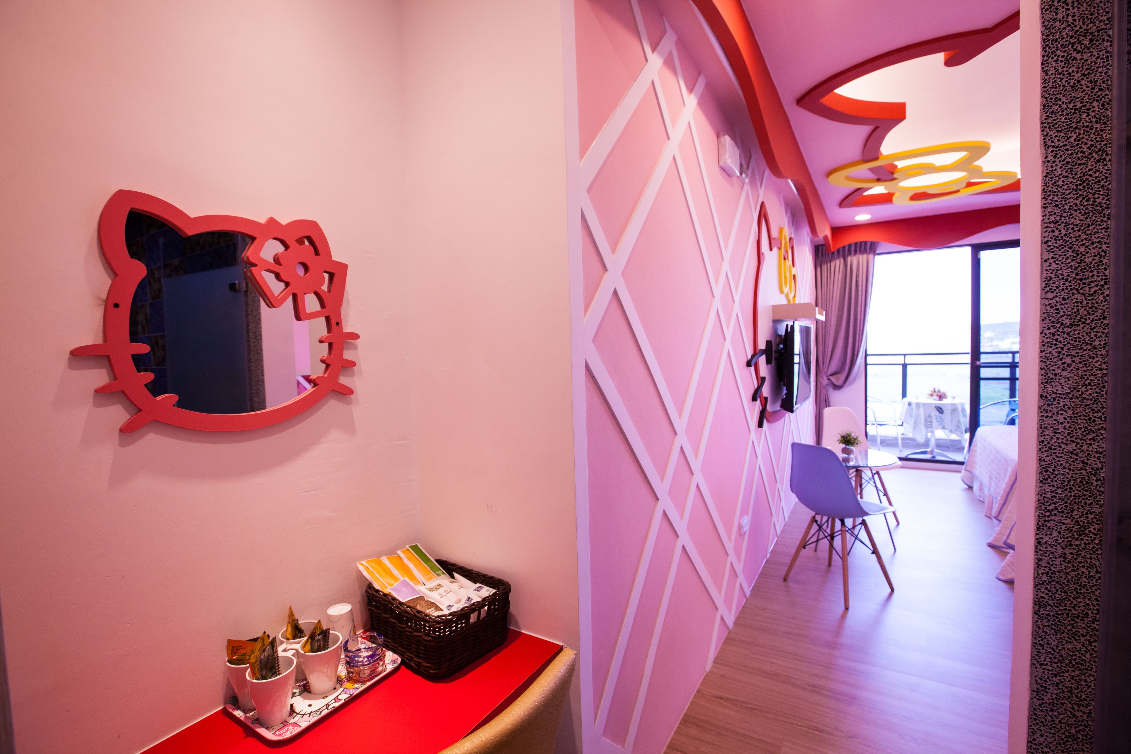 Un pasillo de hotel con un espejo rojo, una cesta de mimbre con artículos de aseo y vistas a un balcón con mesa y sillas.