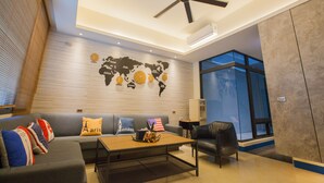 Una sala de estar moderna con un sofá gris, una mesa de centro y un sillón de cuero negro. En la pared hay un mural de un mapa mundial, y un ventilador de techo.
