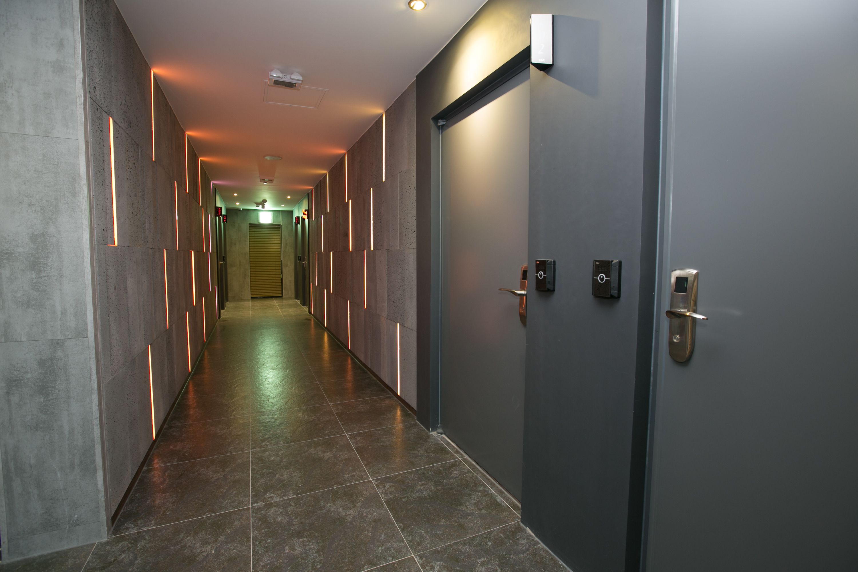 A hallway with a modern design, featuring a long corridor with a tiled floor and walls with embedded lighting. There are doors on the right side with electronic locks.