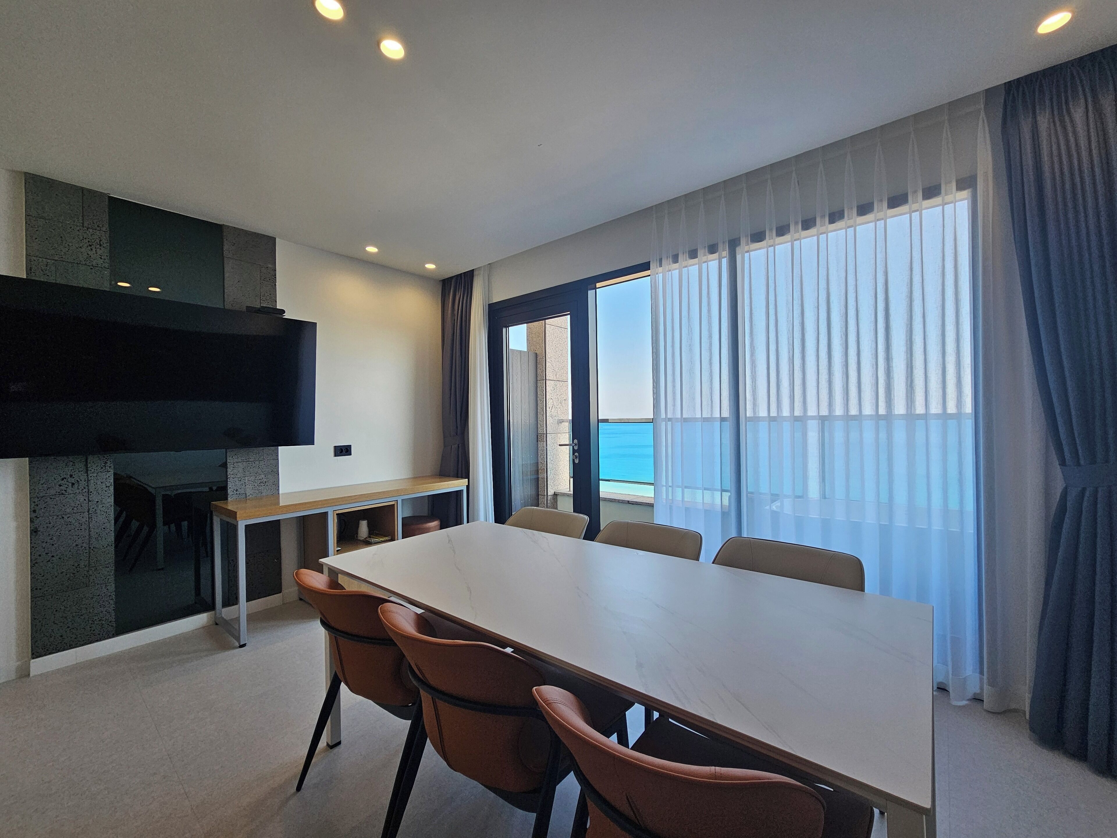 A modern room with a large table, chairs, a flat-screen TV, and a view of the sea through large windows.