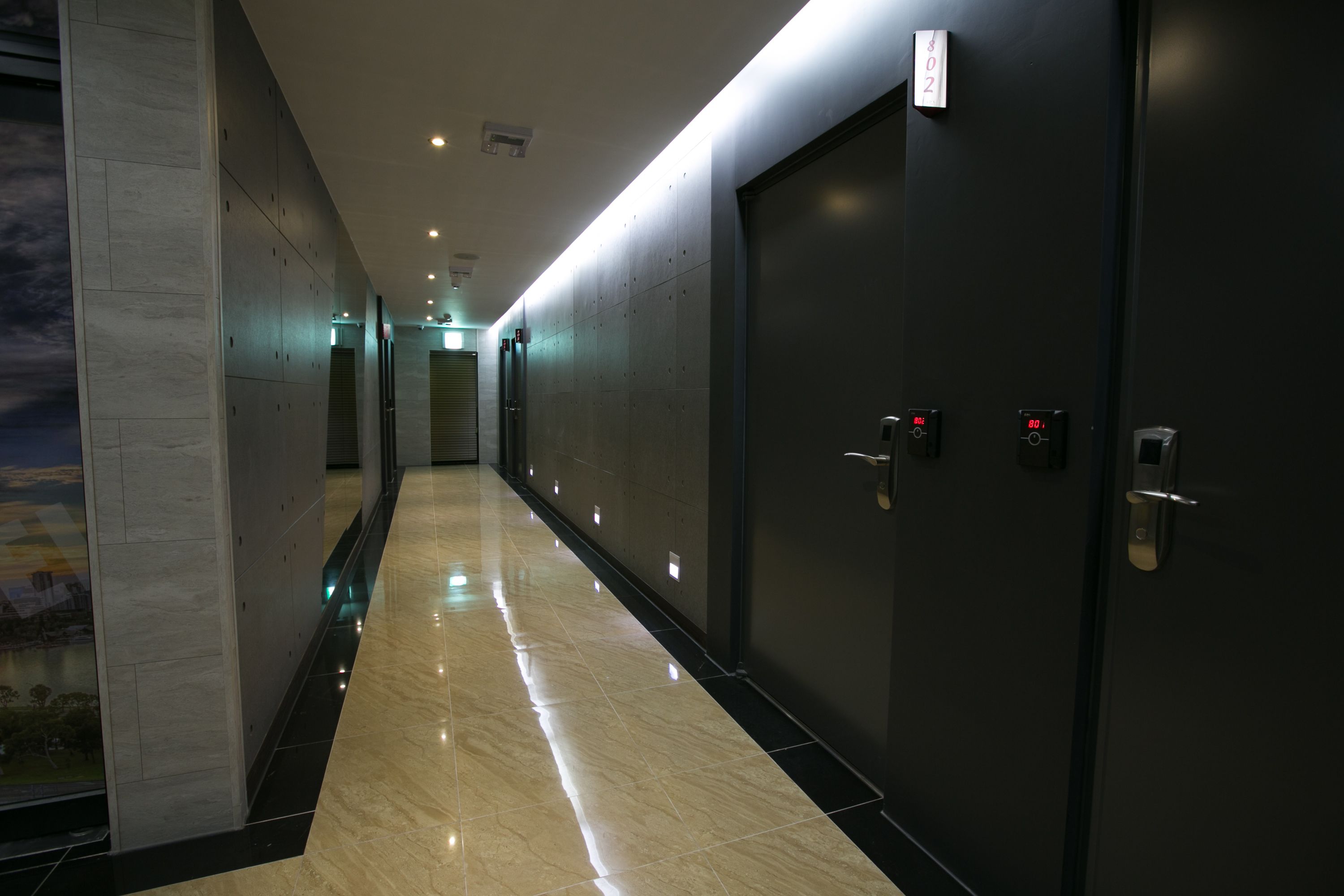 Un pasillo moderno de hotel con piso brillante, paredes oscuras y una puerta grande con cerradura digital.