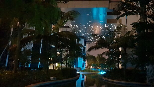 Een nachtzicht van een hotel met een reflecterend zwembad, palmbomen en een gebouw met verlichte ramen.