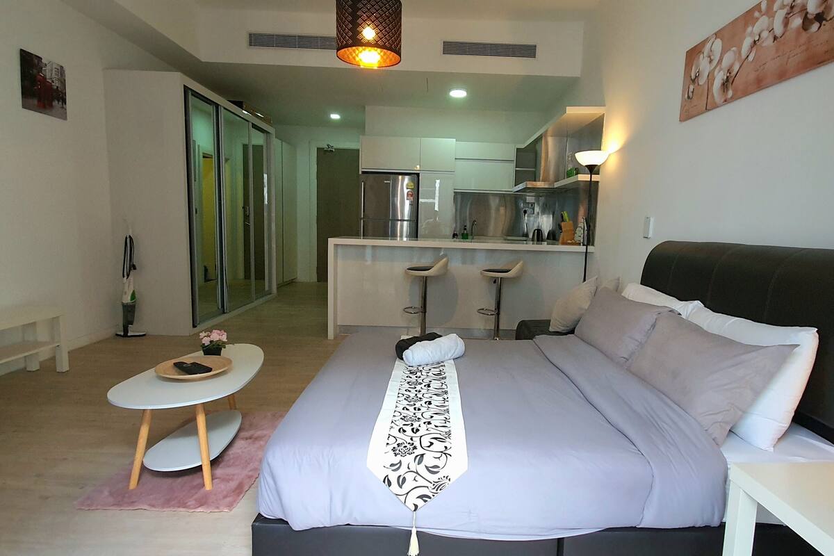 Une chambre d’hôtel moderne avec un grand lit, un bureau, une chaise, une télévision et, au fond, une kitchenette.