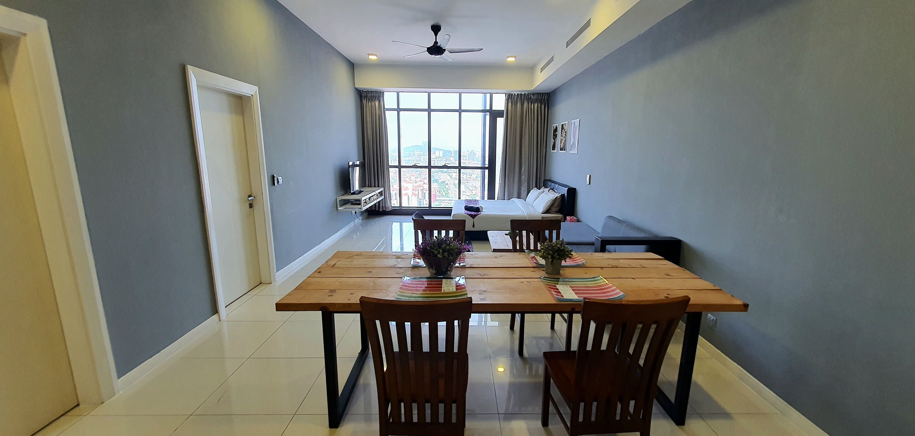 A hotel room with a dining area featuring a wooden table and chairs, a bed with a view of the city, and a ceiling fan.