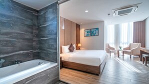 A modern hotel room with a large bed, a separate bathroom with a bathtub, a desk with chairs, and artwork on the wall. - Greenery Hotel (Da Nang)