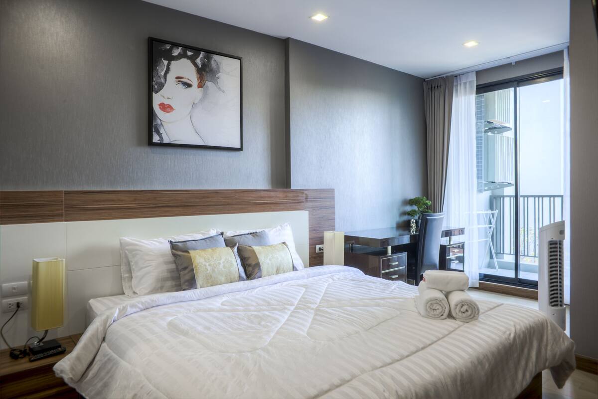 A modern hotel room with a large bed, a desk, a chair, a potted plant, and a view of the outside.